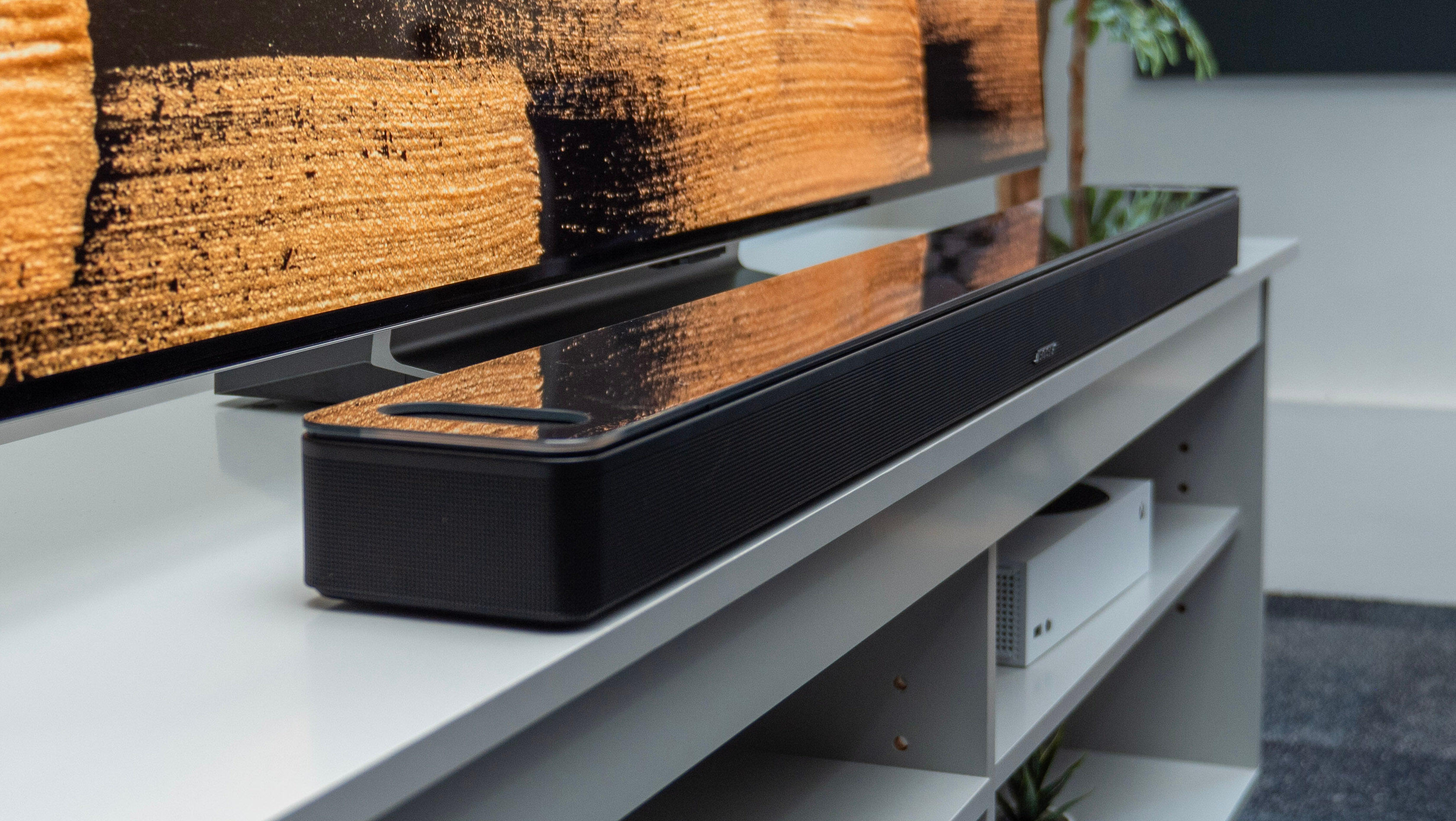 A black Bose Smart Ultra Soundbar sits just in front of a Television on a light grey TV stand. 