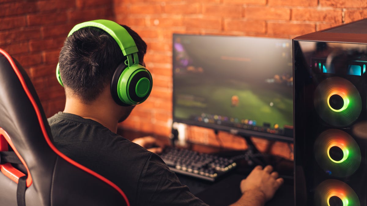 Man with green headphones sits next to his gaming PC and monitor playing World of Warcraft