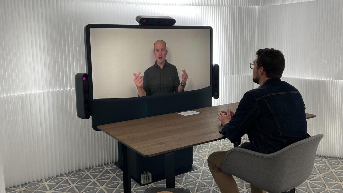 A man sits at a desk in a white hotel room facing a Project Starline prototype: a TV screen ringed by three pods carrying six large cameras, microphones and two speakers.