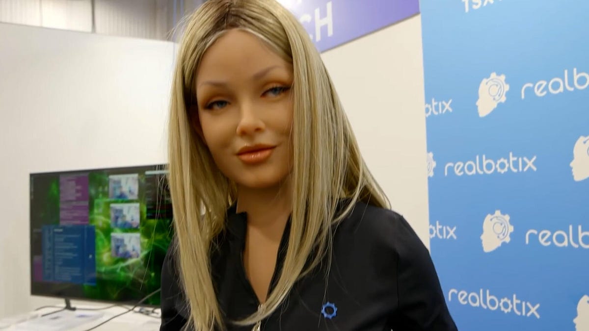 A blonde female robot wearing a black track suit, with a TV screen in the background