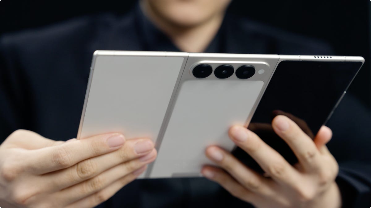 A person holds a tri-fold phone in its fully open state. We see the back, showing the three panels, three cameras, and a front screen.
