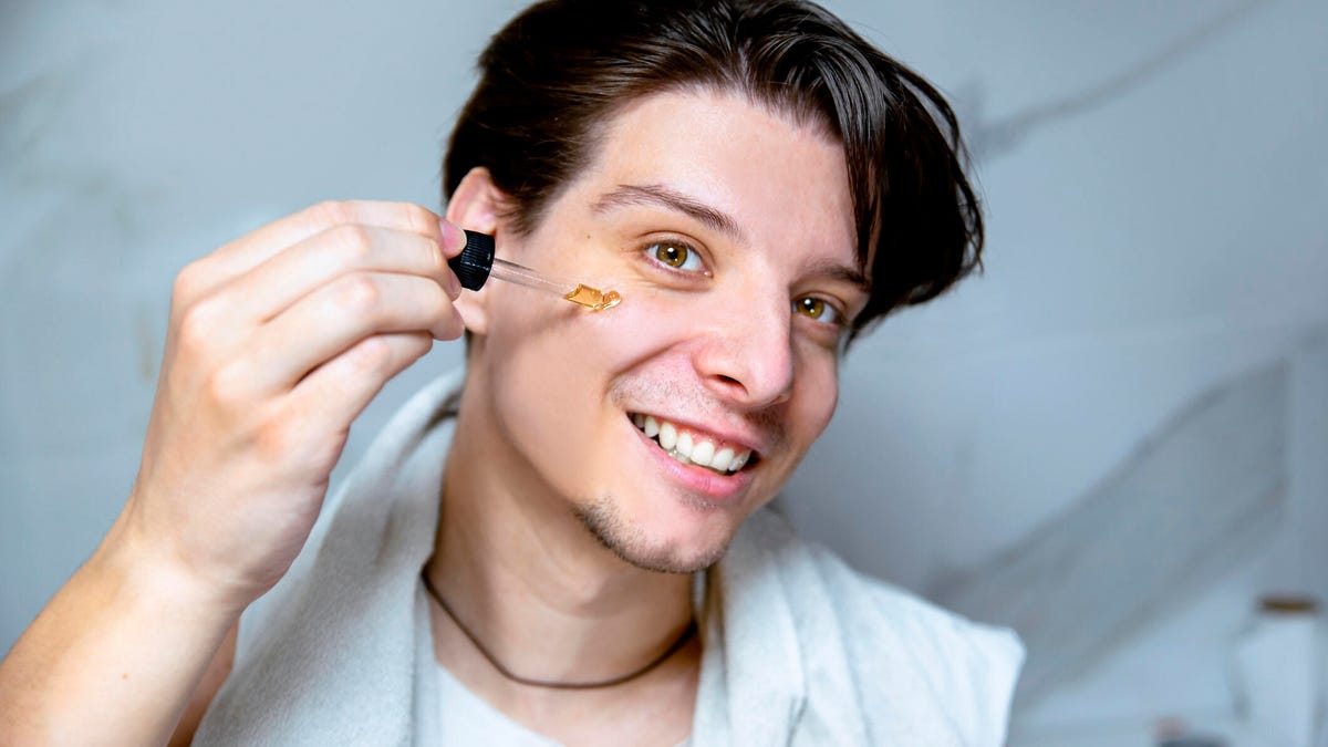 A person with short brown hair and brown eyes using a dropper to applying a yellowish liquid on their face.