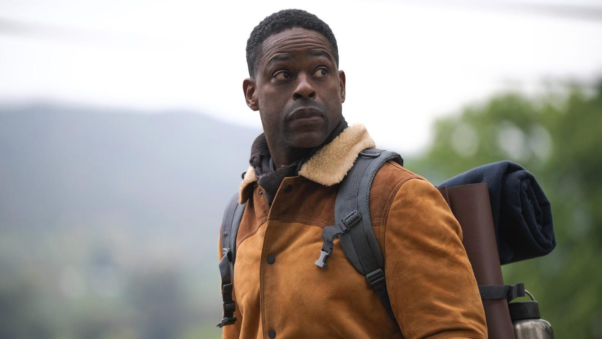 A Black man in a brown jacket wears a backpack and sleeping bag outdoors.