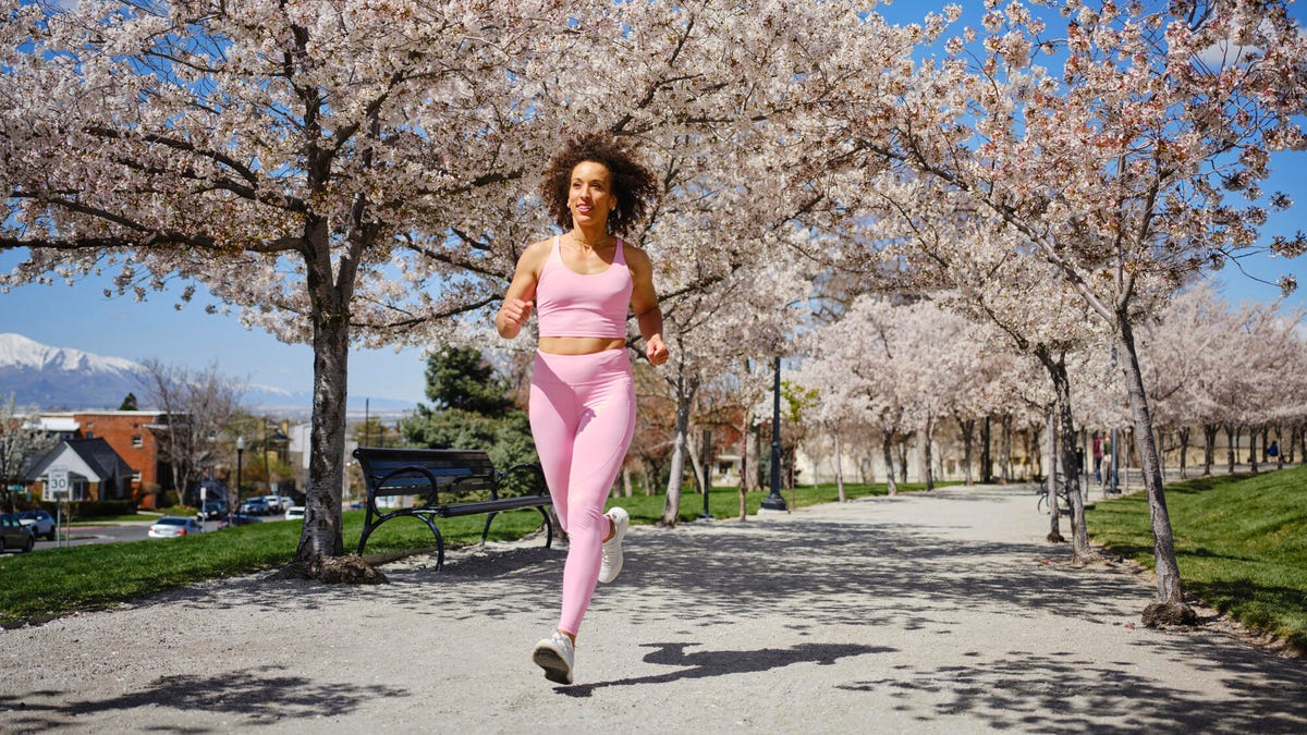 A person running in a light pink sports bra and legging set under cherry blossoms on a pavement path in a park.