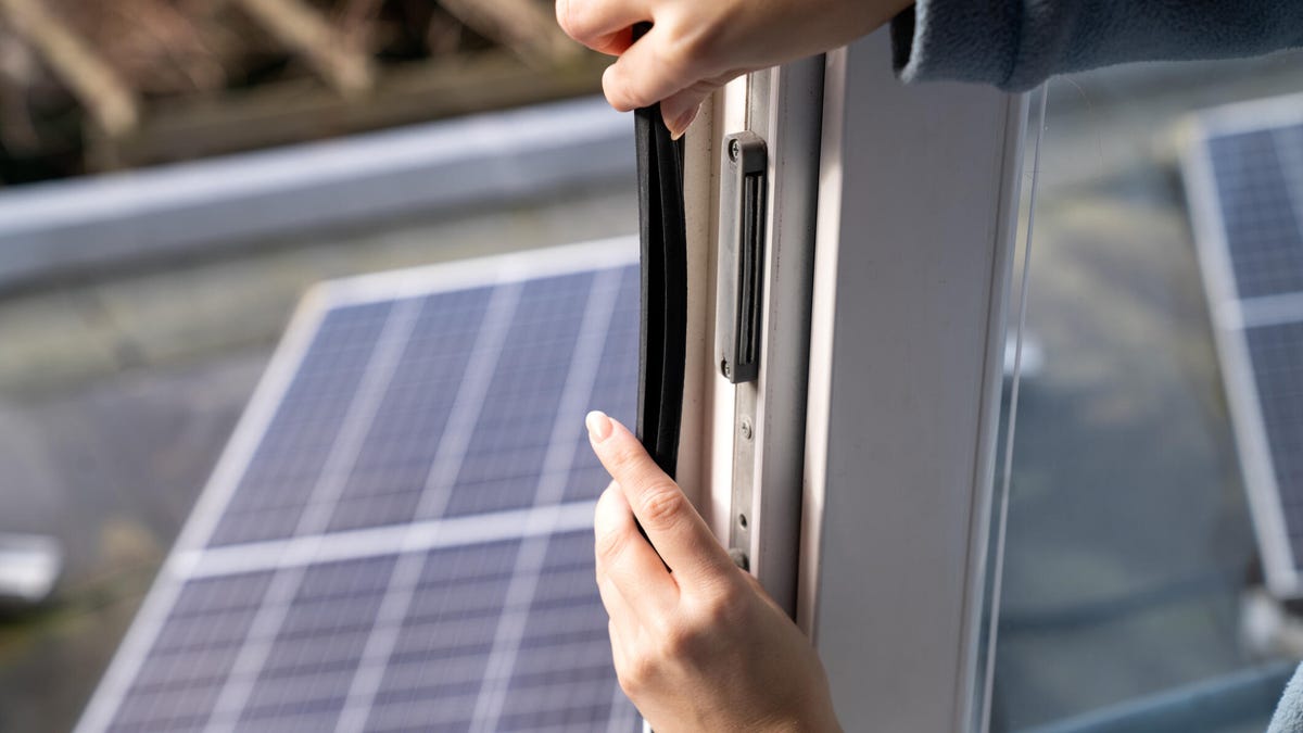 A person applies weatherstripping to a window with a solar panel in the background.