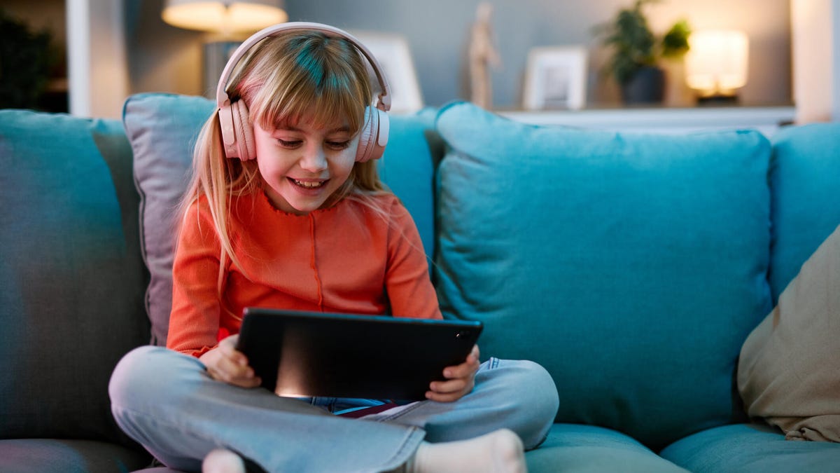 A little girl in an orange top and with pink headphones on her head sits cross-legged on a blue sofa, holding a tablet and smiling