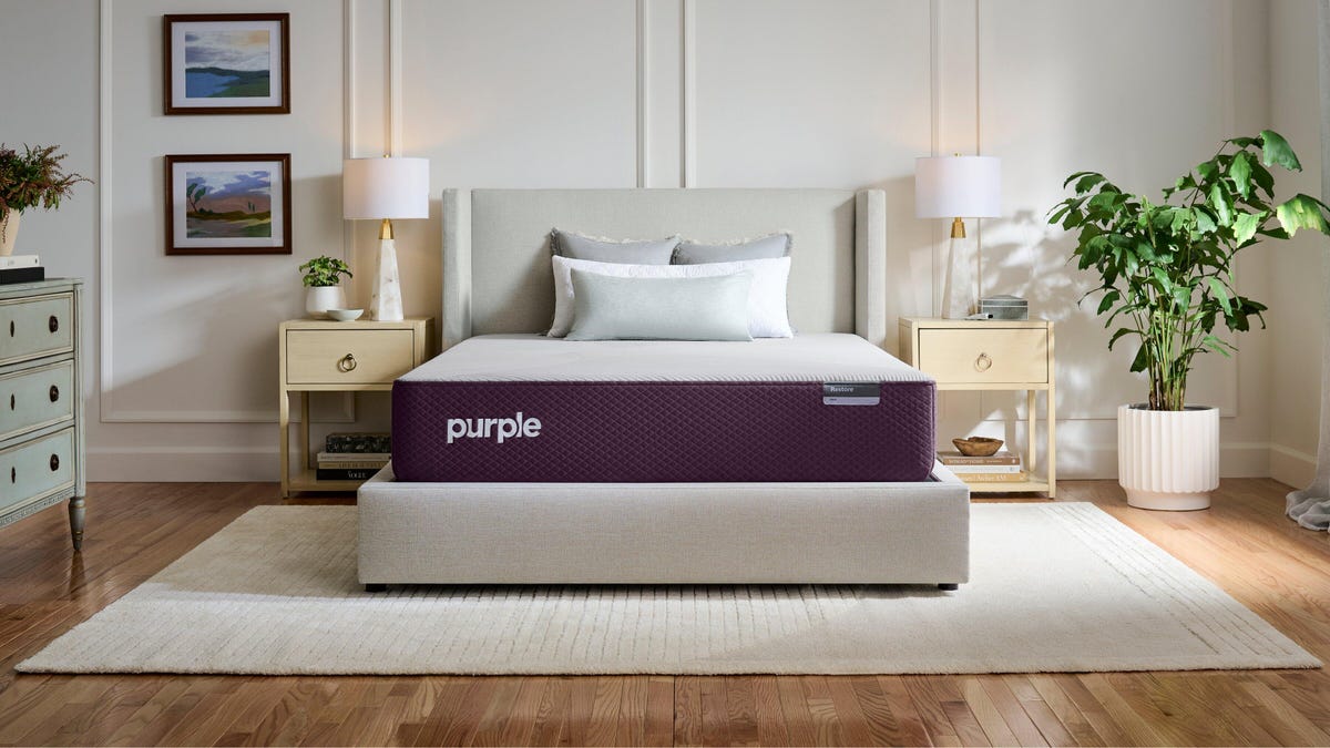 A Purple Restore mattress in a well-lit bedroom with two nightstands and a large plant.