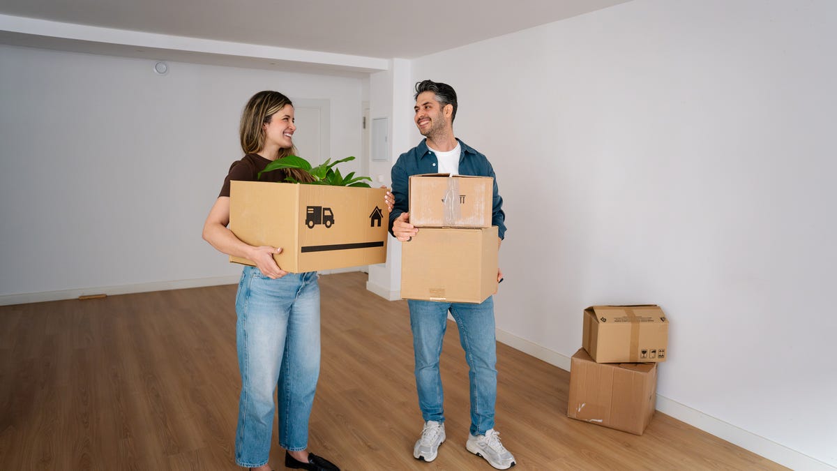 Two people hold moving boxes and smile in an empty room.