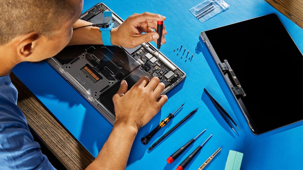 A man bent over a MacBook with the cover removed. He's got a screwdriver poised over the logic board, and the laptop surrounded by neatly organized tools and screws on a blue static mat.