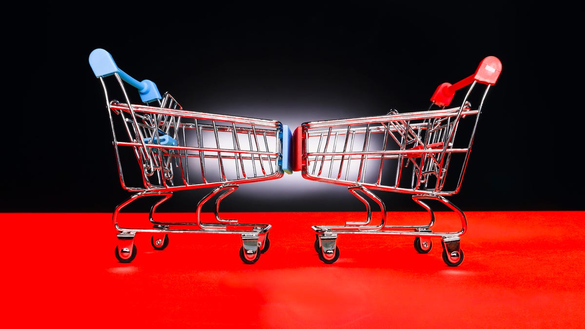 two shopping carts face each other head to head, one with a blue hand and the other red, in front of a black and red background