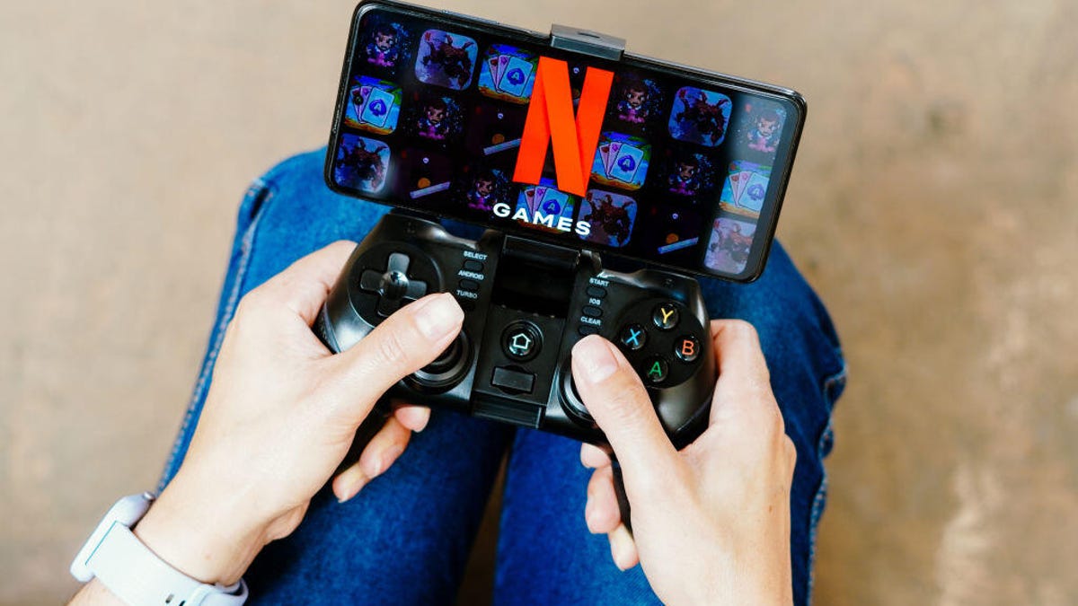 A person holding a gaming controller connected to a smartphone. The smartphone display shows the Netflix games logo