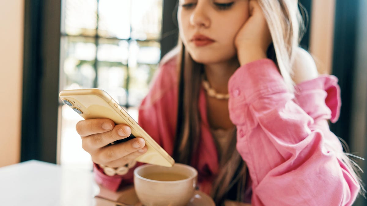 A young woman in a pink shirt checks her phone, with a cup of coffee in front of her