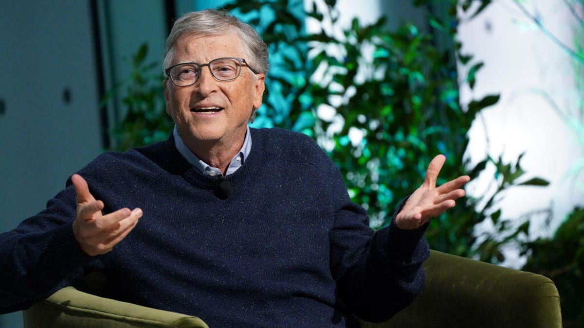 Bill Gates wearing a blue sweater and seated in a chair, gesturing with his hands