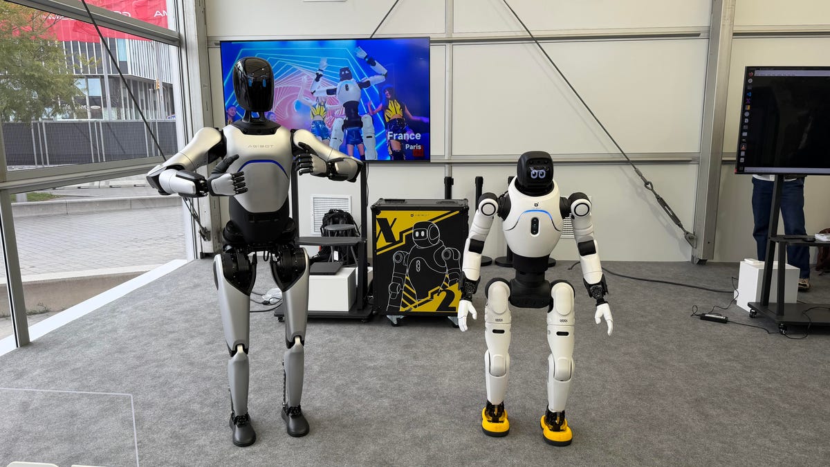 Two white humanoid-styled robots stand side by side in what looks like the inside of an office building. The robot on the left is taller and has its arms lifted outward.