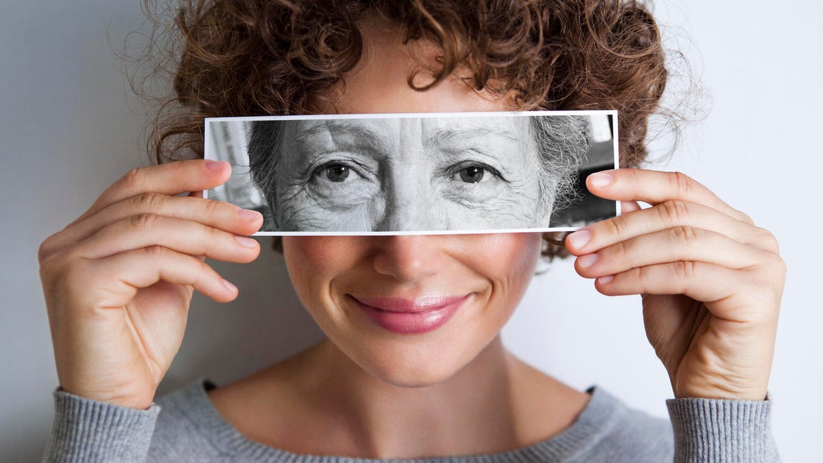 A person with curly brown short hair holding a horizontal black-and-white photo of an older person's eyes over their own eyes.