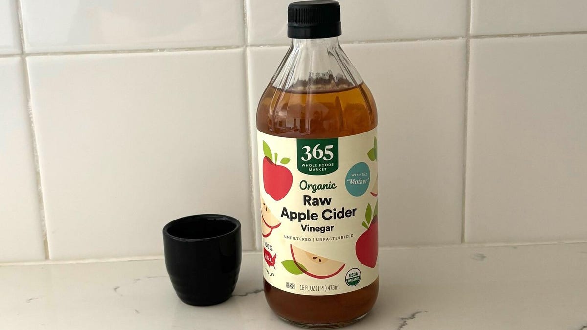 A bottle of Whole Foods 365 raw apple cider vinegar next to a tiny black cup on a marble kitchen counter.