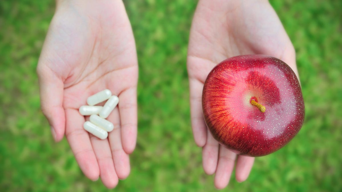 A close-up photo of a person holding a red apple in one hand and white pills in the other over a grassy background.