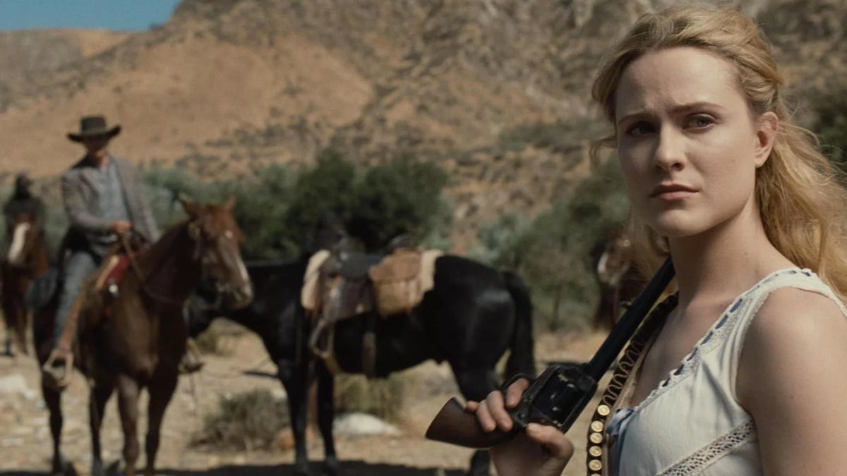 Delores, played by Evan Rachel Wood, stares toward the camera, with two horses (one with a male rider) in the background