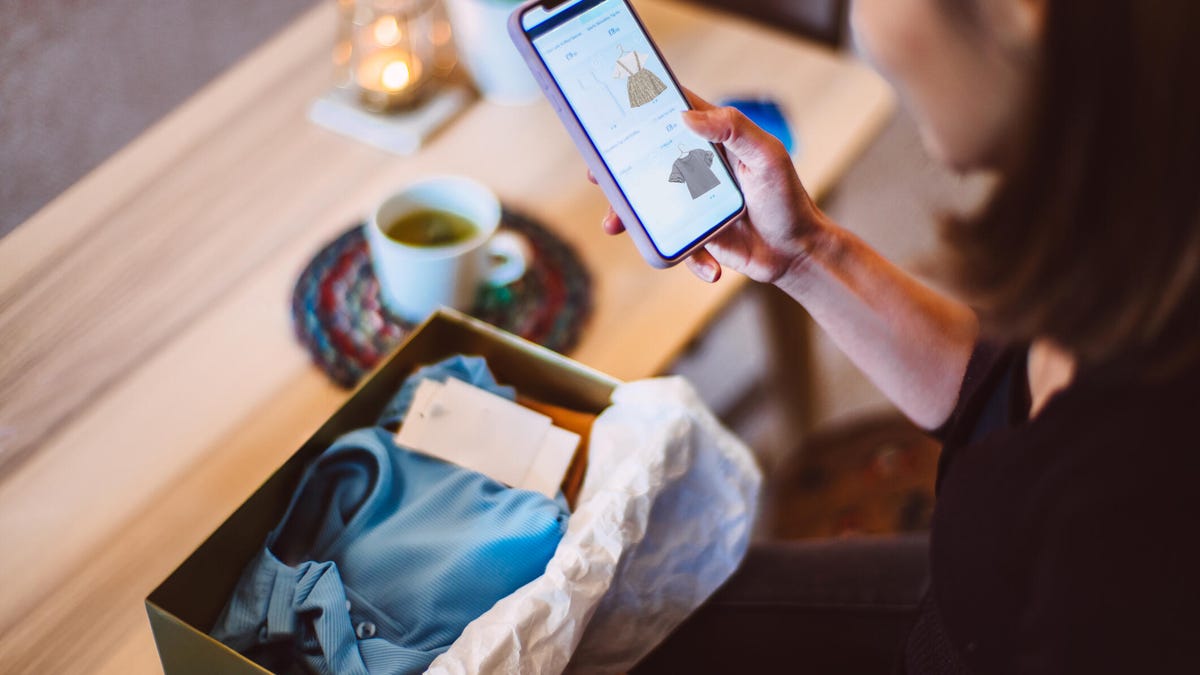 A woman holds a smartphone open to a shopping site while also looking at a box of clothing