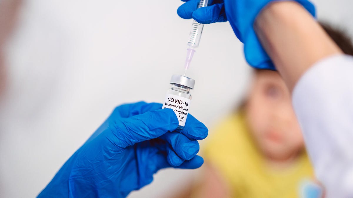 Hands wearing blue surgical gloves prepare a COVID-19 vaccine while a young child looks on