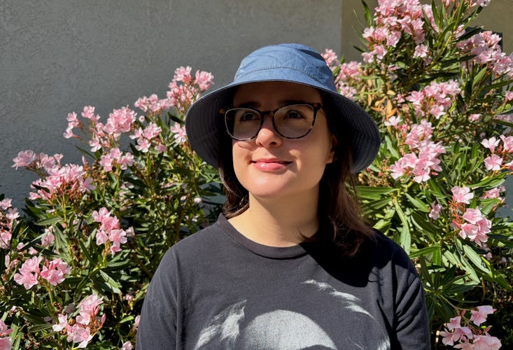 A person with medium-length brown hair wearing the Bondi Bucket Hat in blue in front of a bush of pink flowers outside.