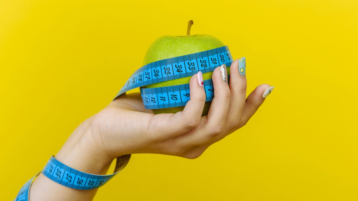 Hand holding an apple wrapped in measuring tape against a yellow backdrop