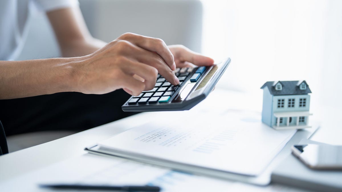 A person holds a calculator over a stack of papers with a small model of a house on the table.