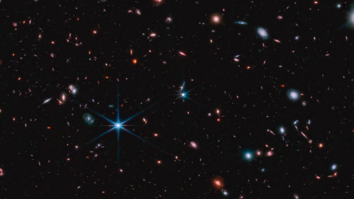 A dark background shows lots of galaxies, some bright, some spiral, some dim, but all in various colors.
