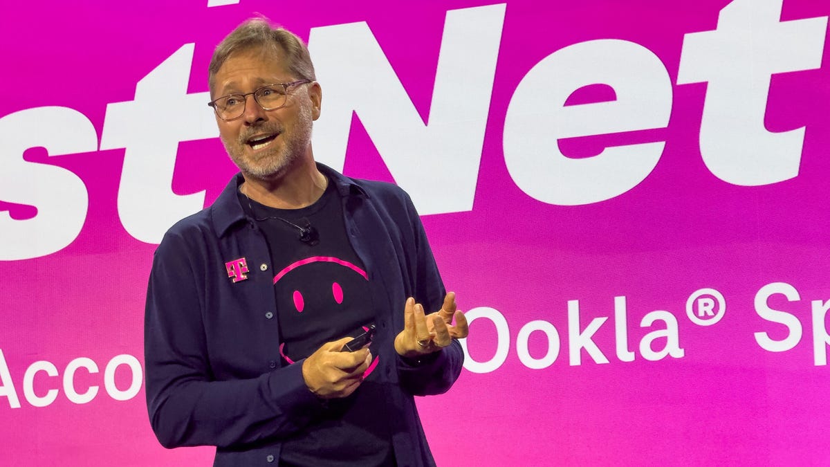 A man in dark clothing and a shirt with a purple smiley face gestures on a stage against the backdrop of a large magenta-colored screen.