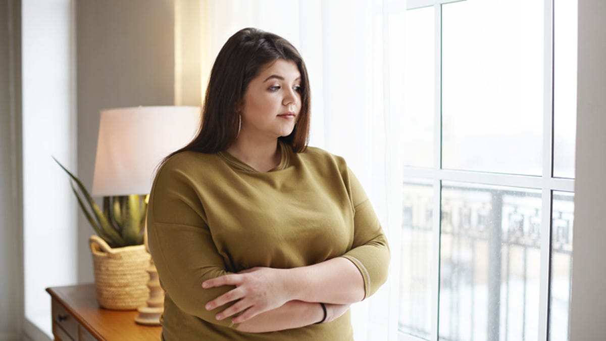 Brunette woman standing in front of a window with her arms crossed over her stomach.
