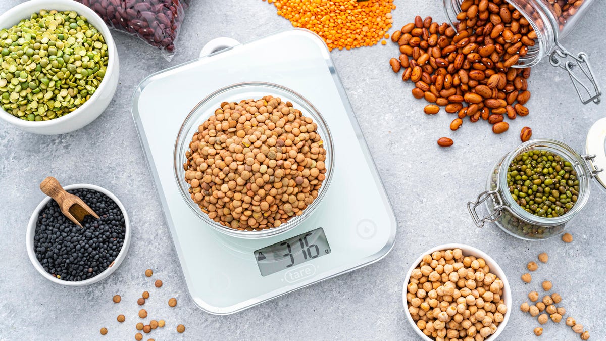 Overhead view of a bowl filled with dried brown lentils placed on a modern kitchen scale. Bowls, bags and containers filled with different legumes are around the kitchen scale and complete the composition.