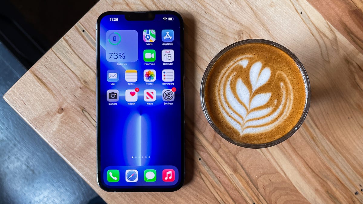 An iPhone 13 sits on a wood table next to a cup of coffee