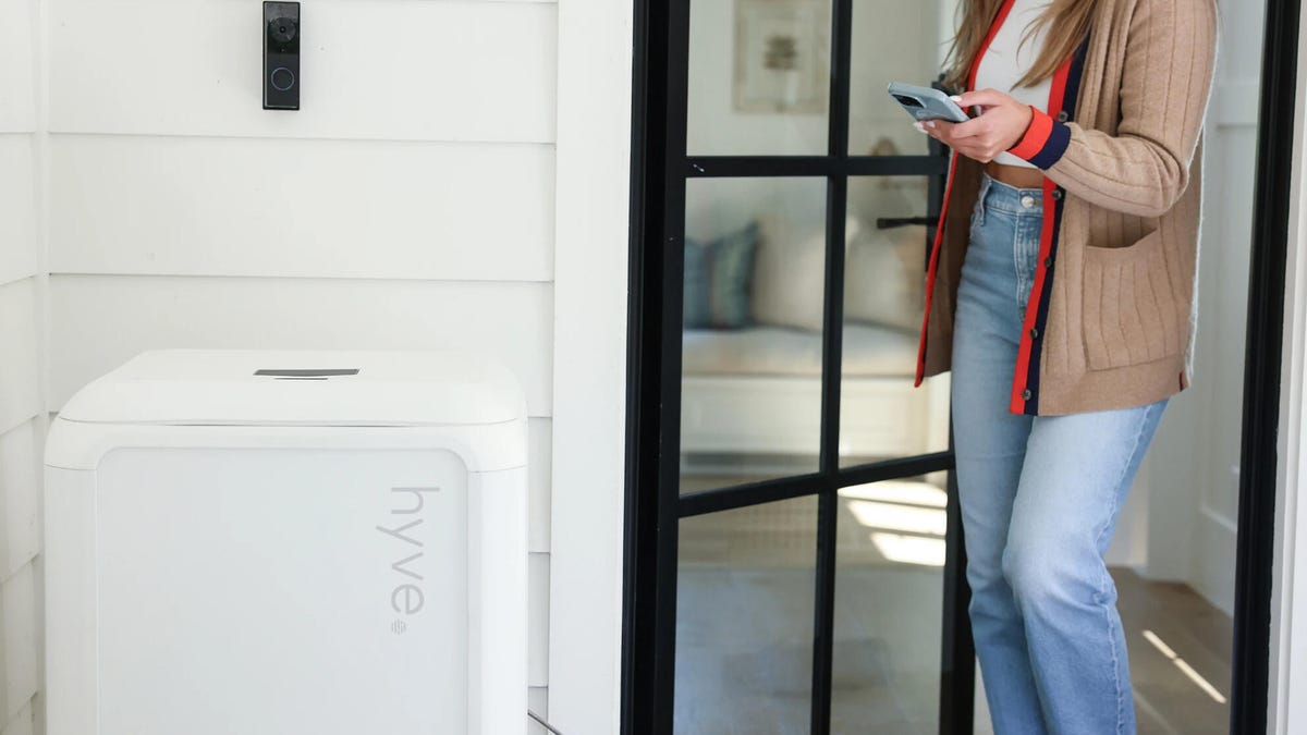 A woman approaches a white Hyve box by a white wall with her phone out.