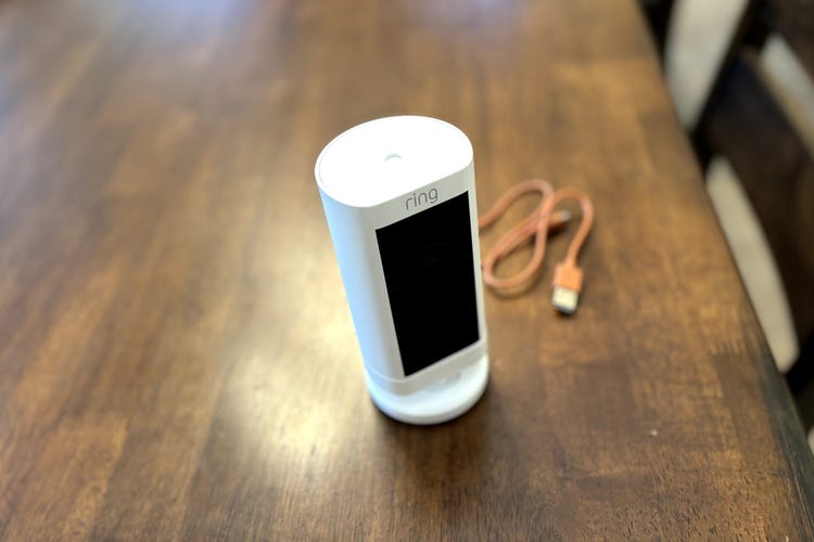 A Ring Outdoor Cam Pro on a wooden table with its unplugged charging table.