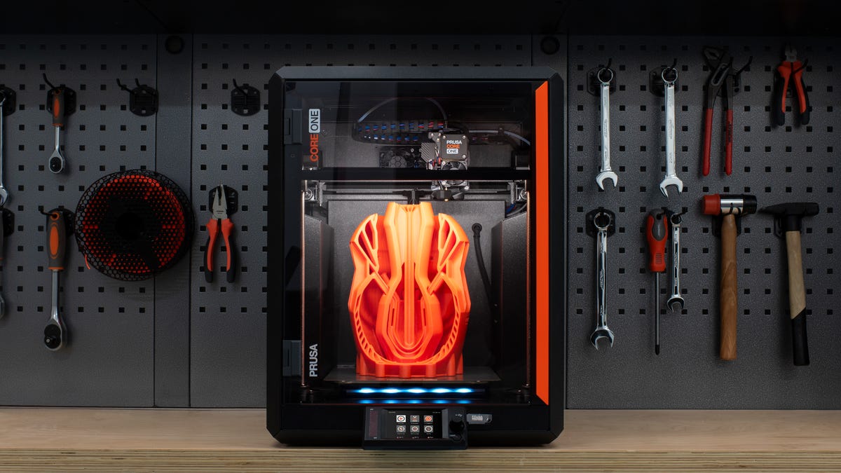 A black 3D printer in front of a black peg board with tools hanging on it.