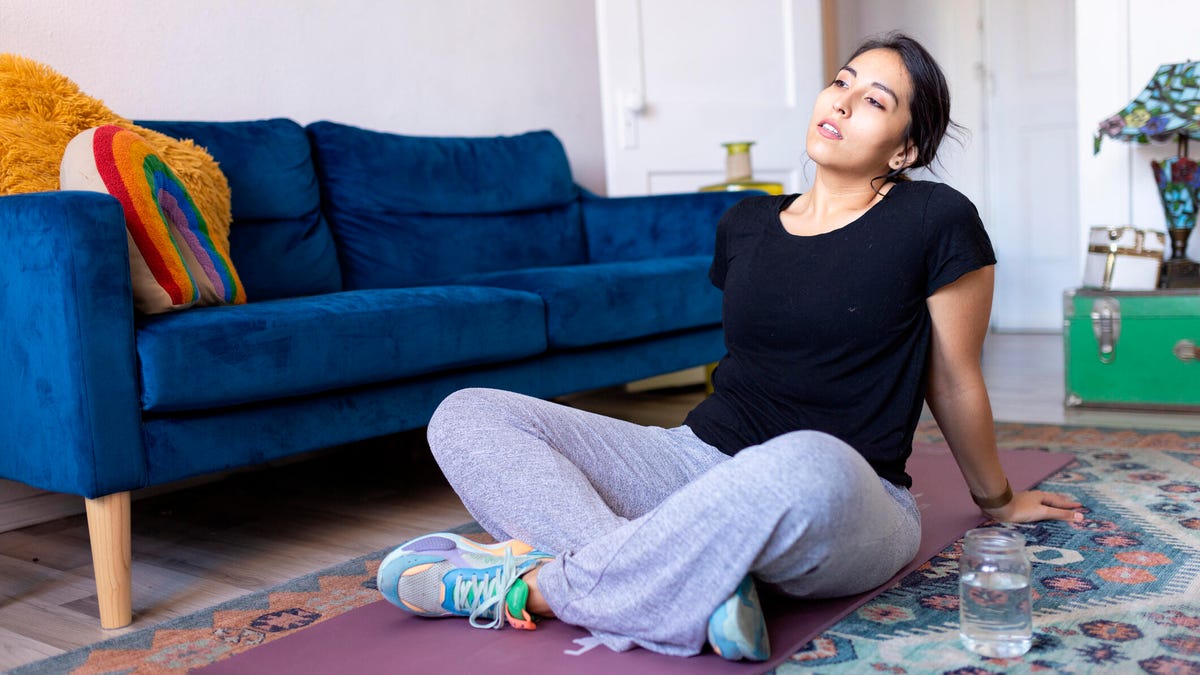 A person with pulled-back black hair, a black tee and grey sweatpants sitting on a yoga mat looking tired in a living room.