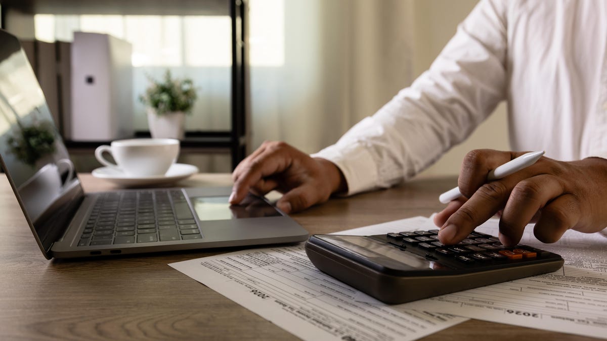 a person in a white shirt uses a laptop with their right hand and a calculator with their right hand. the calculator is sitting on top of a bunch of tax forms, and a white coffee cup sits to the right of the laptop