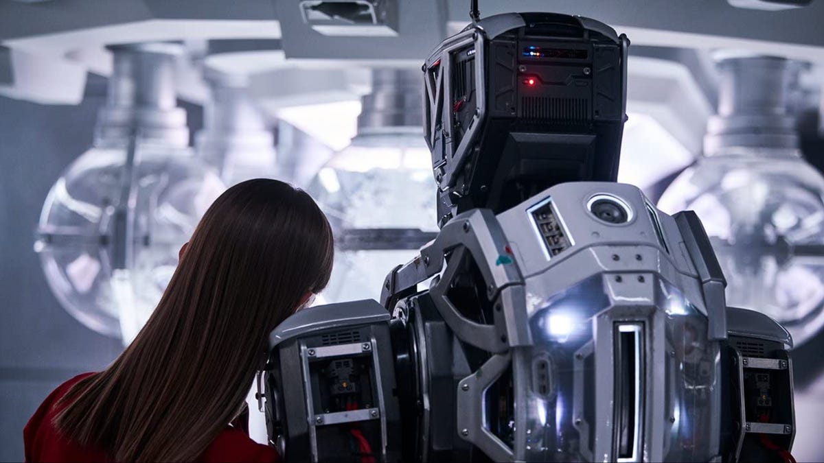 A robot on the right stands taller than a girl on the left. The girl is resting her shoulder on the robot. Their backs are faces us.