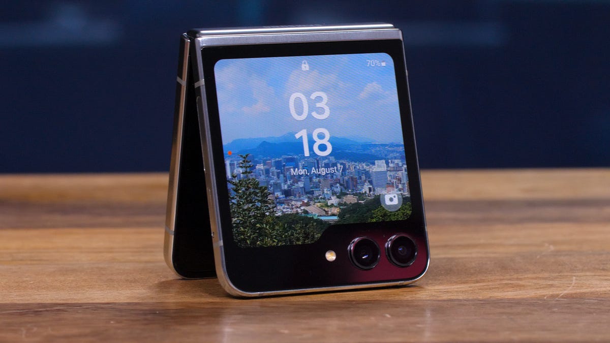 A folding phone "tented" upright on a table with its exterior screen lit and facing the reader, showing the time and a pretty landscape background.