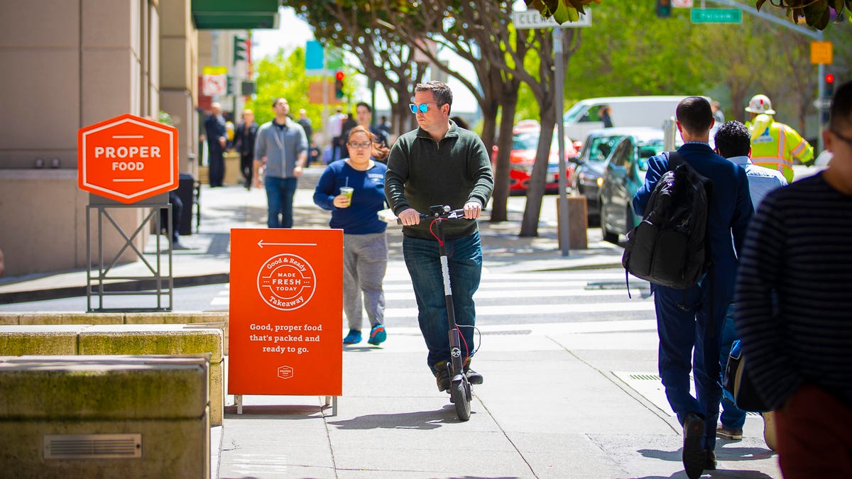 Man riding an e-scooter along a sidewalk, without a helmet.
