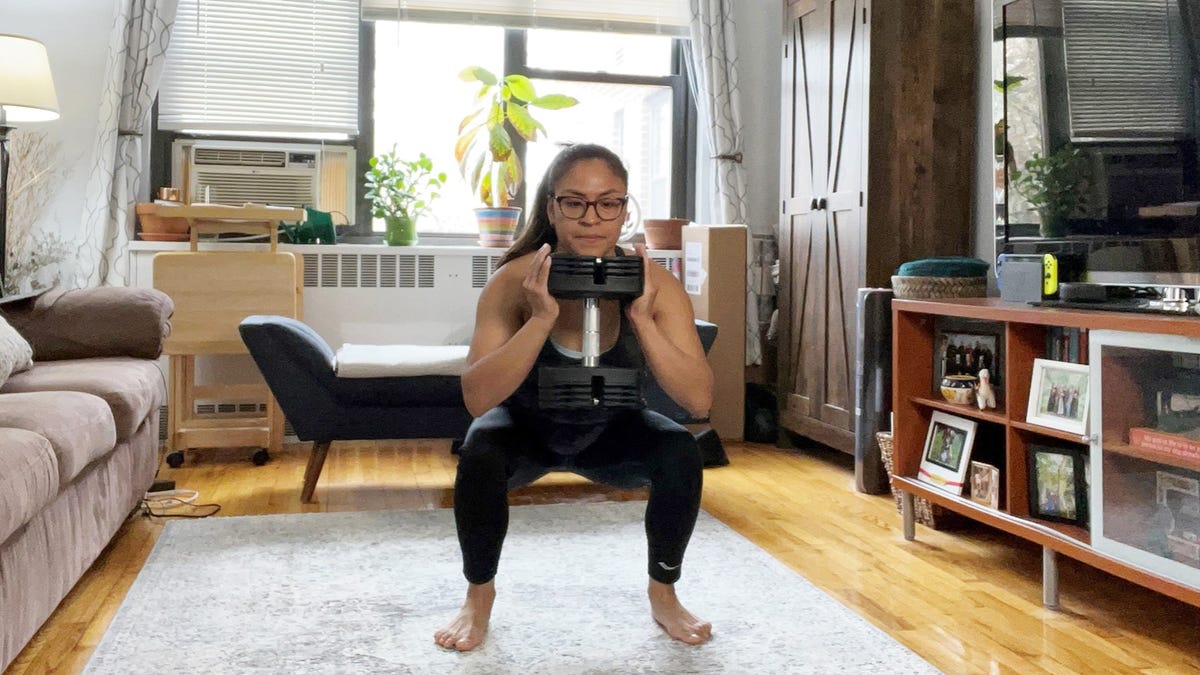 Giselle, a latina woman in workout gear and glasses, squats in her living room, holding a heavy-looking dumbbell between two hands under her chin