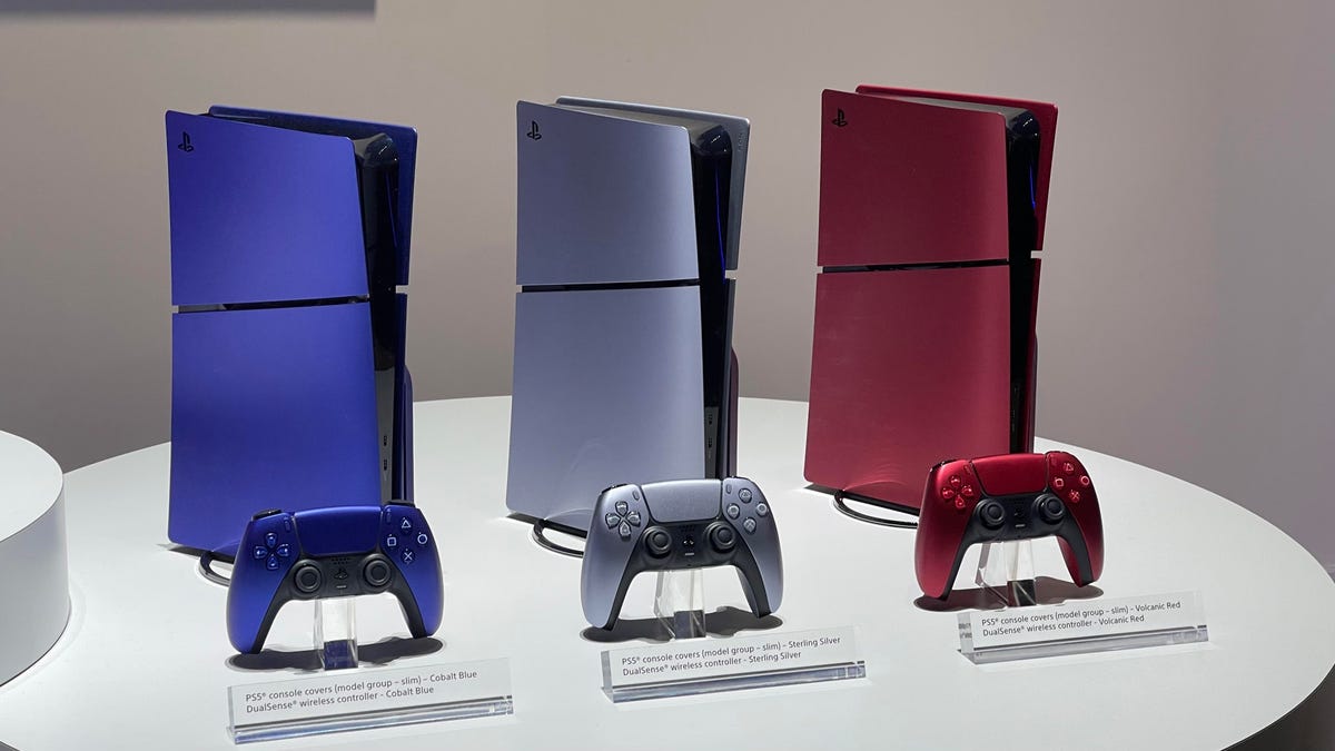 Three PlayStation 5s side by side. One is blue, one is silver and the third is red. PlayStation 5 controllers are in front of each.