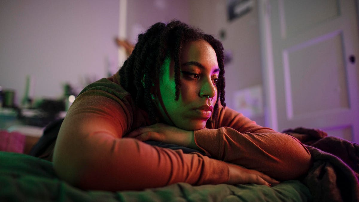 A person lying on a bed, head on crossed arms, a pensive expression on their face