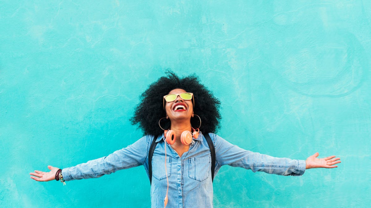 Woman wearing sunglasses standing in front of a blue background with her arms out.