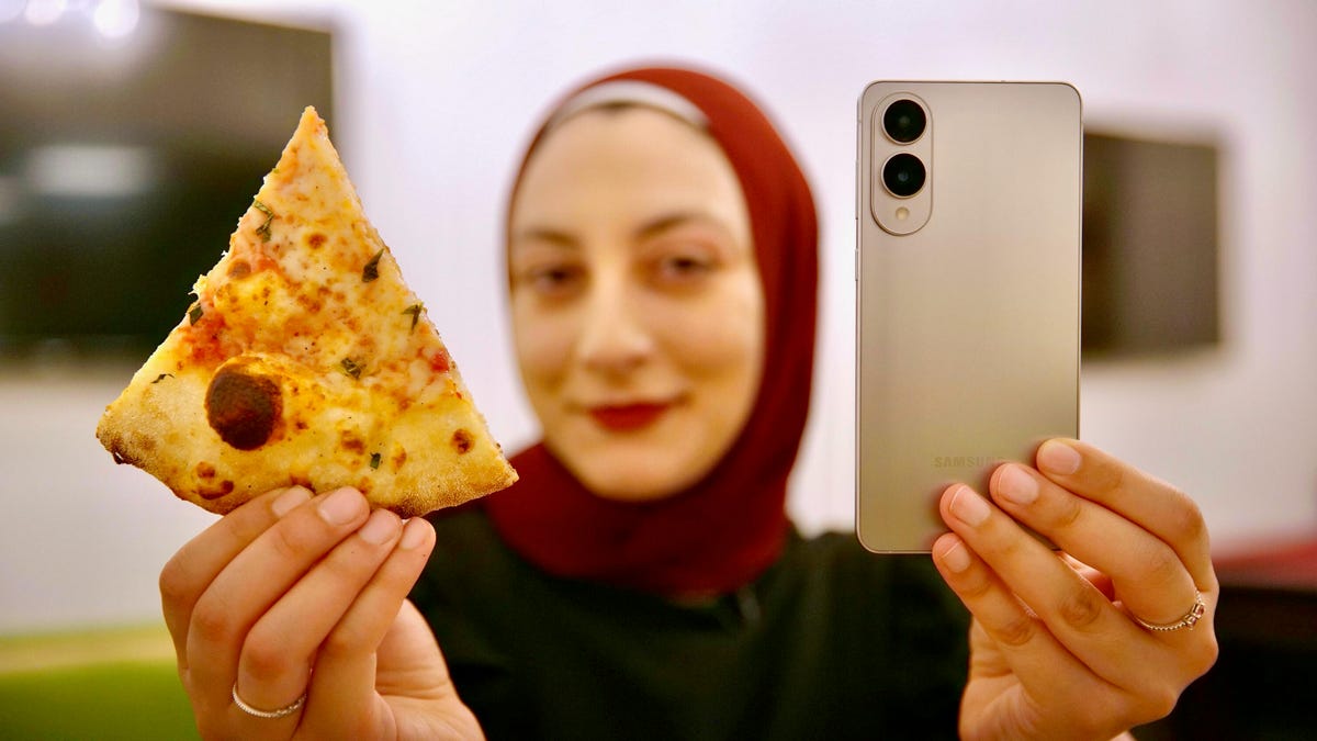 A woman in a red headscarf hold the Galaxy S25 Edge in one hand and a slice of thin-crust pizza in another