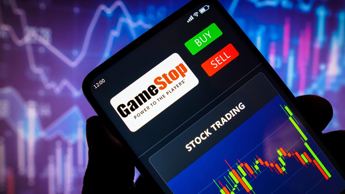 a hand holding a phone showing buttons to buy and sell gamestop stock and a stock chart