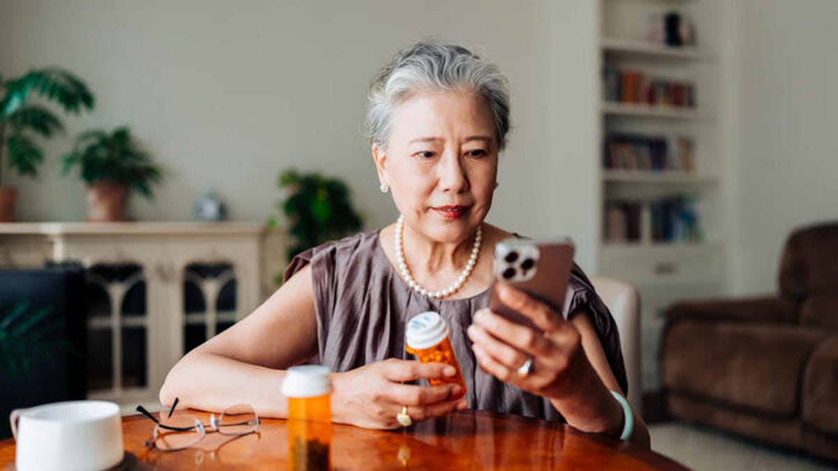 Person sitting at a table holding a pill bottle in one hand and a smartphone in the other health