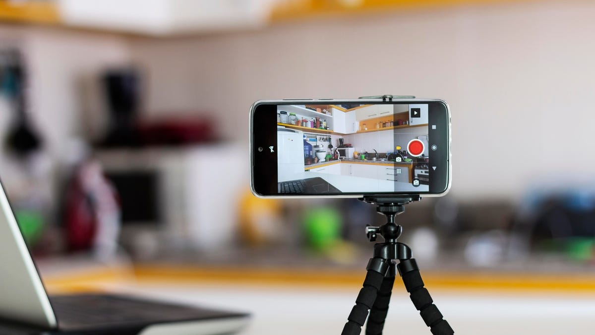 a smartphone is mounted on a tripod next to a laptop on a counter. the camera is facing a kitchen