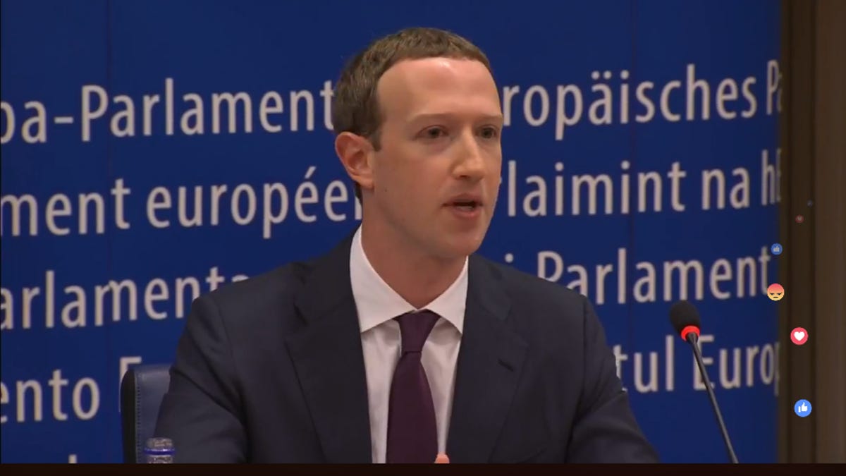 Mark Zuckerberg in suit and tie talks, sitting in front of a blue banner with multi-lingual messaging on it. In the foreground, a heart, angry face, and "like" symbol float in front of the Facebook livestream of the event.