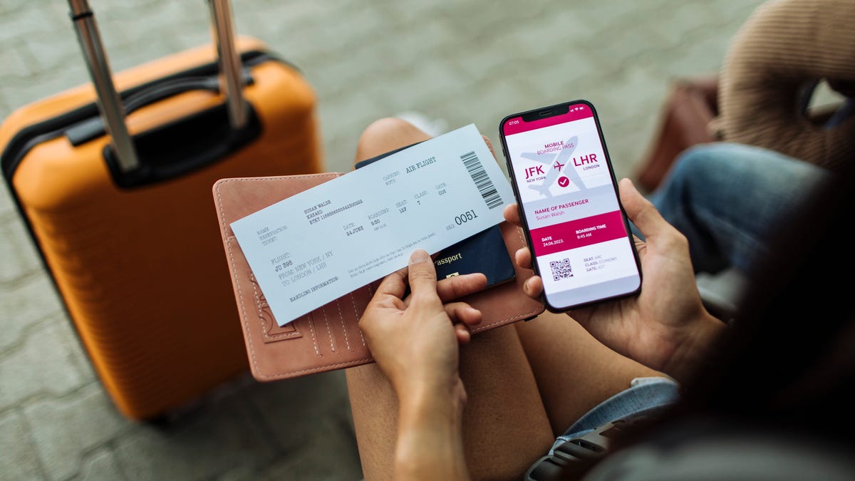 a person holds a smartphone with a flight itinerary showing while also looking at a printed boarding pass as they sit in front of an orange rolling suitcase with its handle extended